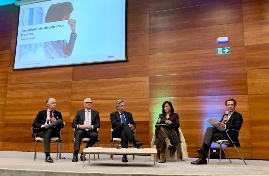 Mesa redonda compuesta por cuatro hombres y una mujer en un auditorio con paredes de madera. Sobre ellos, una gran pantalla proyecta una presentación titulada "Absentismo: del diagnóstico a la acción". El ponente de la derecha interviene con un micrófono y unos apuntes en la mano.