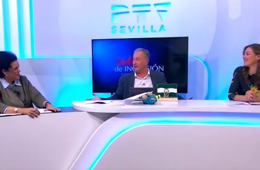 Tres personas en una mesa de debate en el plató de PTV Sevilla. En el centro, un hombre interviene junto a una pequeña bandera de Andalucía, flanqueado por dos mujeres que escuchan la conversación. El fondo es de color blanco y azul brillante con el logotipo de la cadena.
