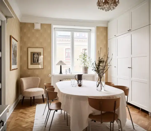 Comedor con papel pintado dorado botánico, mesa ovalada con mantel blanco y sillas de madera con patas metálicas